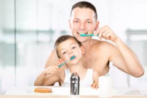 Young father and son brushing their teeth together as they watch in a bathroom mirror.