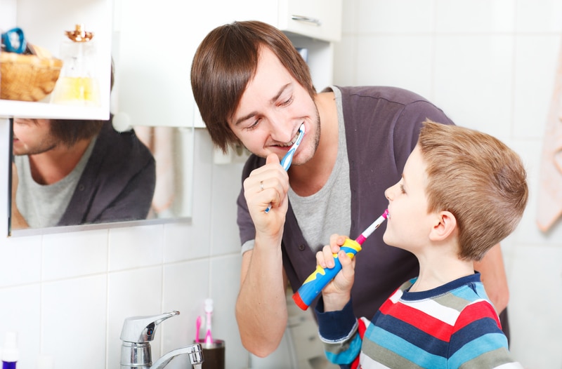 Young adult male brushing his teeth alongside a young boy in a bathroom, presumably they are father and son.