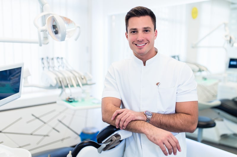 A male dentist that is smiling at the camera and leaning against a dental chair. There are dental tools and equipment in the background.