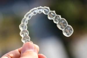A view of a transparent orthodontic aligner that someone is holding up in front of a darker background. 