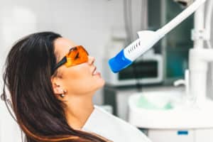 Brunette woman that is wearing protective orange glasses as she is getting a teeth whitening light treatment