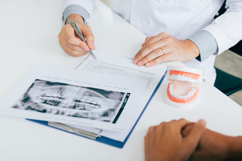 An image of a dentist's hands as he is filling out papers with a patient. There is a dental model of a healthy mouth and dental x-rays with the paperwork.