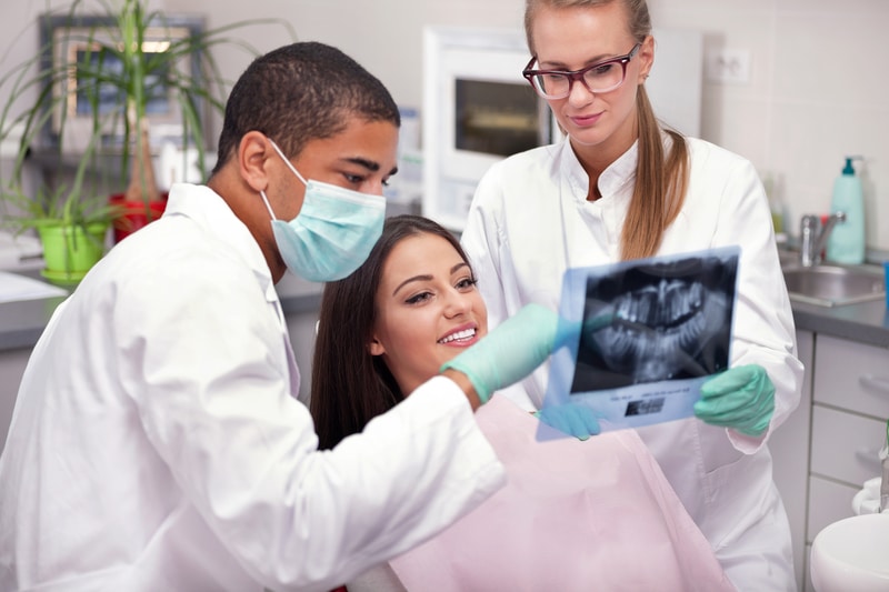 A brunette young adult woman that is in a dental chair going over her dental x-rays with the dentist and hygienist.