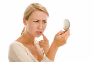 A blonde, young adult woman that is looking in a makeup compact mirror, pointing to a spot near her mouth and making a confused face about what the spot could be.