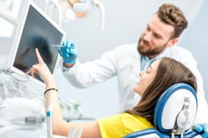 A young adult male dentist that is pointing at an x-ray image in front of his young adult brunette woman patient.