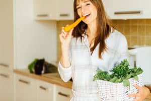 A young adult woman that is eating a carrot with one hand and holding a basket of leafy greens in another arm. 