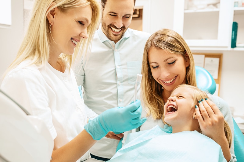 A family of a mother, father and a young daughter that are laughing as a dental assistant is with them. 