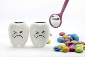 Two tooth models sitting side by side that have frowning faces on their faces. The top of the teeth models are black and decayed and there are pieces of candies behind the teeth.