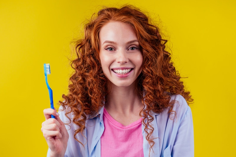 Red-headed woman with curly hair that is smiling straight at the camera holding a toothbrush. She is wearing a pink shirt, lavender sweatshirt and is in front of a mustard-yellow background.