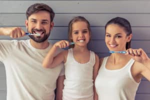 Beautiful parents and their daughter are looking at camera and smiling while brushing teeth