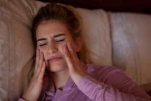 A woman lying on her back in bed holding her jaw