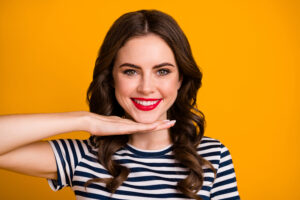 Smiling brunette woman in a striped shirt, holding her hand under her chin to display her new smile after veneers